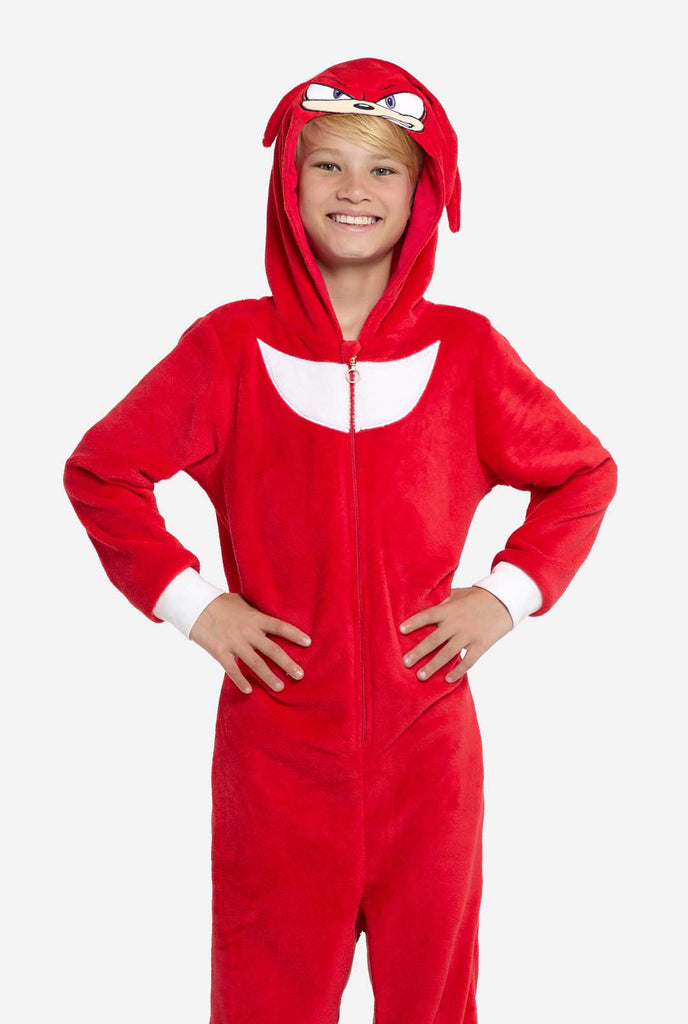 A kid is wearing a bright red Knuckles the Echidna-themed kids onesie from OppoSuits. The fun and cozy onesie features a zip-up front and an attached hood designed to resemble Knuckles' face, complete with expressive eyes and floppy dreadlocks. The suit is primarily red, with a white crescent pattern on the chest, reflecting Knuckles' iconic look from the Sonic the Hedgehog video game series. 