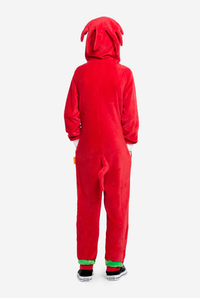 A kid is wearing a bright red Knuckles the Echidna-themed kids onesie from OppoSuits. The fun and cozy onesie features a zip-up front and an attached hood designed to resemble Knuckles' face, complete with expressive eyes and floppy dreadlocks. The suit is primarily red, with a white crescent pattern on the chest, reflecting Knuckles' iconic look from the Sonic the Hedgehog video game series. 