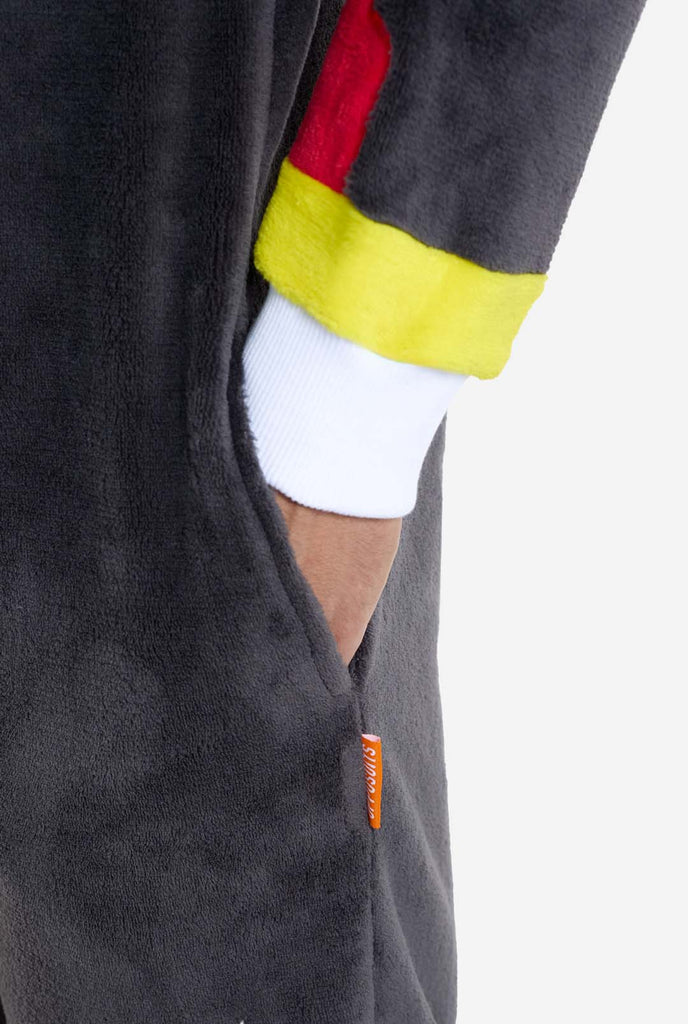 A man wearing a plush black onesie inspired by the popular hedgehog character Shadow, featuring spiky red and black accents on the hood, a white chest emblem, and yellow wrist details. The onesie is made from soft fleece material, offering comfort and a playful design for cosplay, lounging, or themed events.  Perfect for fans of gaming or animated characters. Sleeve close up with hand in the pocket. 