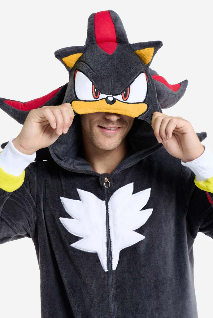 A man wearing a plush black onesie inspired by the popular hedgehog character Shadow, featuring spiky red and black accents on the hood, a white chest emblem, and yellow wrist details. The onesie is made from soft fleece material, offering comfort and a playful design for cosplay, lounging, or themed events. The person smiles while adjusting the hood, showcasing the character's recognizable face and bold design. Perfect for fans of gaming or animated characters. Close up.