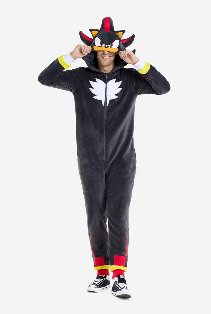 A man wearing a plush black onesie inspired by the popular hedgehog character Shadow, featuring spiky red and black accents on the hood, a white chest emblem, and yellow wrist details. The onesie is made from soft fleece material, offering comfort and a playful design for cosplay, lounging, or themed events. The person smiles while adjusting the hood, showcasing the character's recognizable face and bold design. Perfect for fans of gaming or animated characters. Full body image.