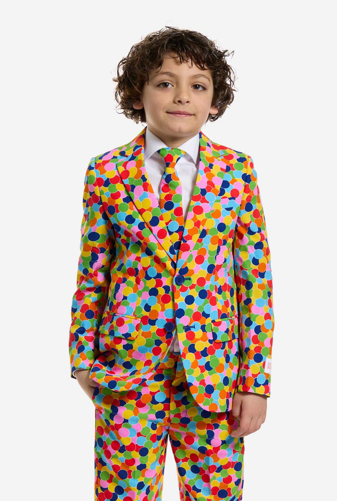 In this image, a kid is wearing a boys’ suit with all-over multicolor confetti print and white shirt. The suit has a bright pattern with circles in various colors including orange, yellow, pink, green, blue, and red.