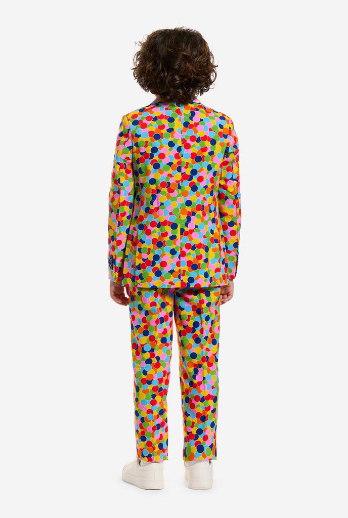 In this image, a kid is wearing a boys’ suit with all-over multicolor confetti print and white shirt. The suit has a bright pattern with circles in various colors including orange, yellow, pink, green, blue, and red. View from the back.