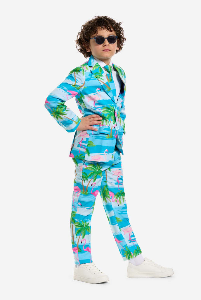 In this image, a kid is wearing a light blue boys’ suit with a tropical print and a white shirt. The suit has a blue print resembling water and pink flamingos and green palm trees. Kid wearing sunglasses.