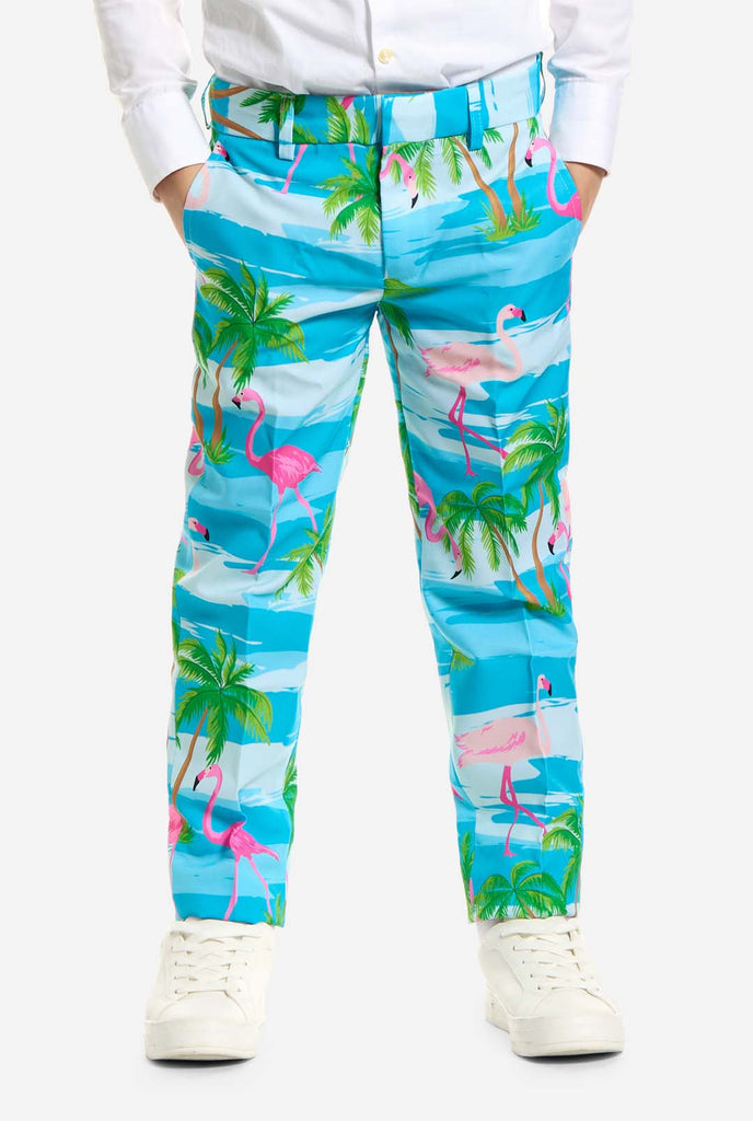 In this image, a kid is wearing a light blue boys’ suit with a tropical print and a white shirt. The suit has a blue print resembling water and pink flamingos and green palm trees. Pants close up.