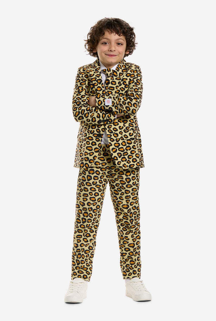 In this image, a kid is wearing a boys’ suit with all-over panther print and white shirt. The suit is beige with brown and black spotted panther print. Suits consists of jacket, pants and clip-on tie. Standing with crossed arms.