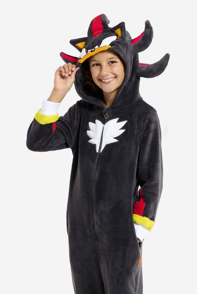 A kid wearing a plush black kids onesie inspired by the popular hedgehog character Shadow, featuring spiky red and black accents on the hood, a white chest emblem, and yellow wrist details. The onesie is made from soft fleece material, offering comfort and a playful design for cosplay, lounging, or themed events. The person smiles while adjusting the hood, showcasing the character's recognizable face and bold design. Perfect for fans of gaming or animated characters.