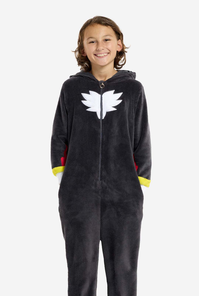 A kid wearing a plush black kids onesie inspired by the popular hedgehog character Shadow, featuring spiky red and black accents on the hood, a white chest emblem, and yellow wrist details. The onesie is made from soft fleece material, offering comfort and a playful design for cosplay, lounging, or themed events. The person smiles while the hood is lying on the back. Perfect for fans of gaming or animated characters.