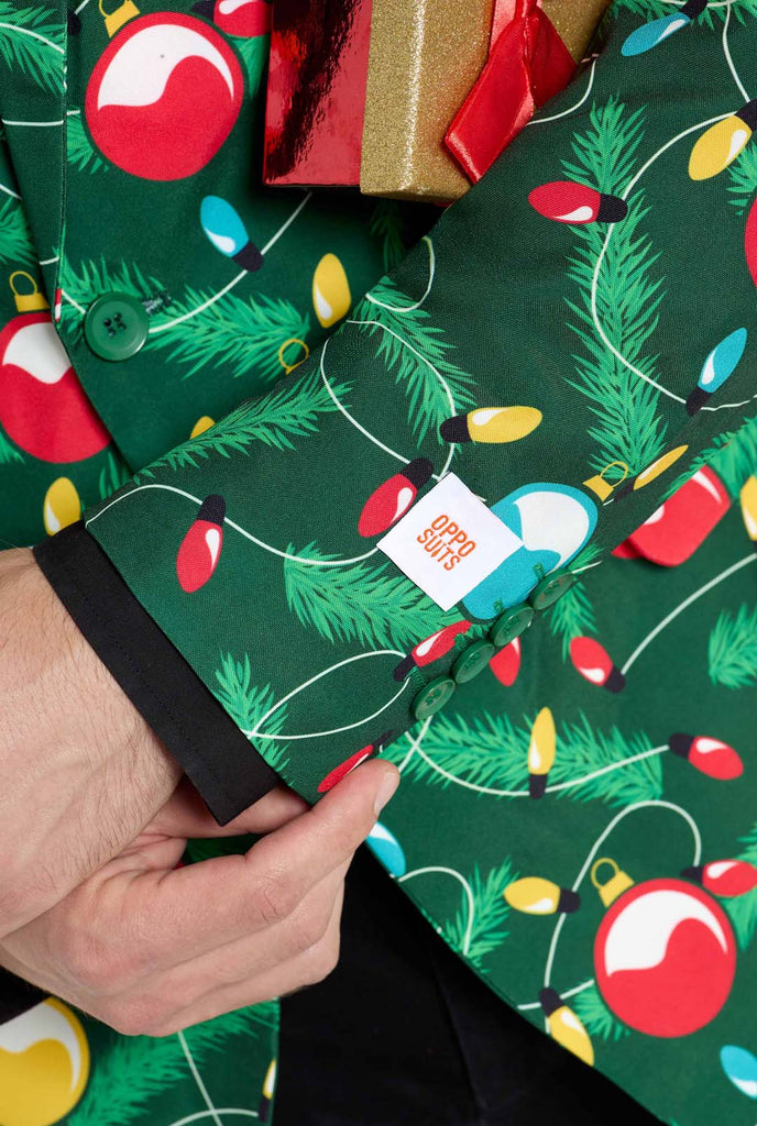 Man wearing green Christmas blazer for men with decorated Christmas tree print. Man also wearing a black pants and black men's shirt. Sleeve close up.