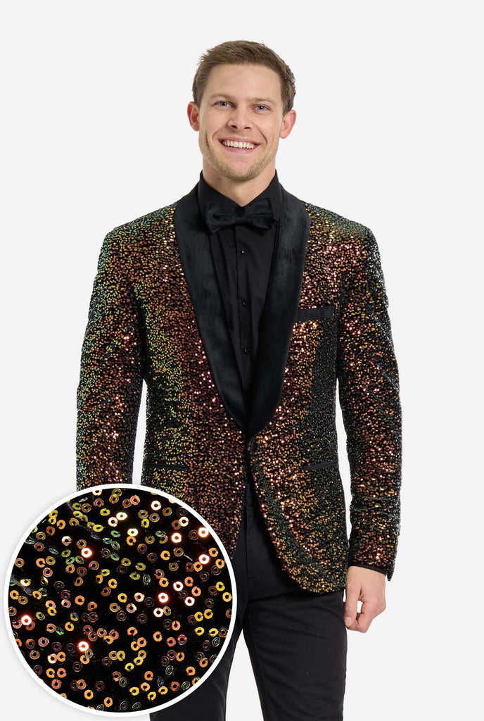 A man is standing and smiling, dressed in a formal, eye-catching outfit. He is wearing a black dress shirt with a black bow tie and a blazer adorned with shimmering amber-colored sequins that reflect light in various shades of gold, copper, and green. The blazer has a satin black shawl collar and front pockets. He pairs this bold jacket with black dress pants, creating a contrast between the sparkly top and the sleek bottom. The background is plain, emphasizing the dazzling effect of the sequined blazer.