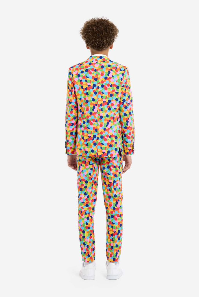 In this image, a kid is wearing a colorful, polka-dotted suit. The suit has a bright pattern with circles in various colors including orange, yellow, pink, green, blue, and red, arranged in a confetti-like style. View from the back.