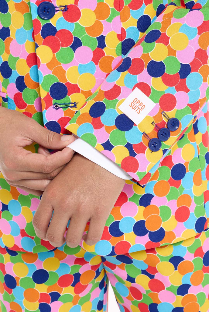 In this image, a kid is wearing a colorful, polka-dotted suit. The suit has a bright pattern with circles in various colors including orange, yellow, pink, green, blue, and red, arranged in a confetti-like style. Sleeve close up.