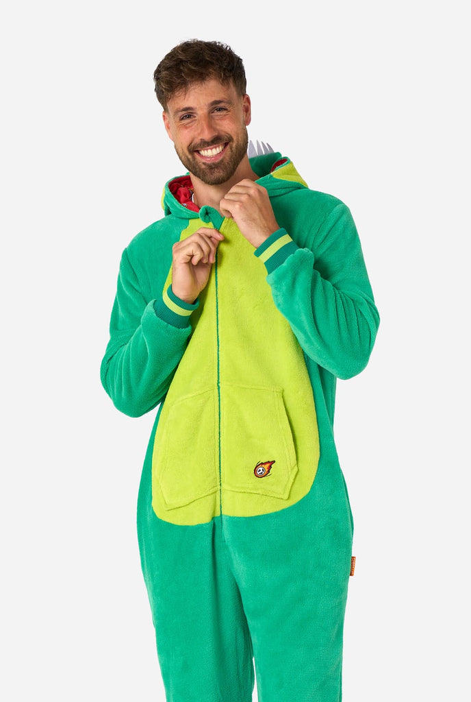 A man is wearing a green dinosaur adult onesie featuring playful hood details and cozy, soft material, perfect for lounging or costume events. He is smiling towards the camera and the hood is down on his back.