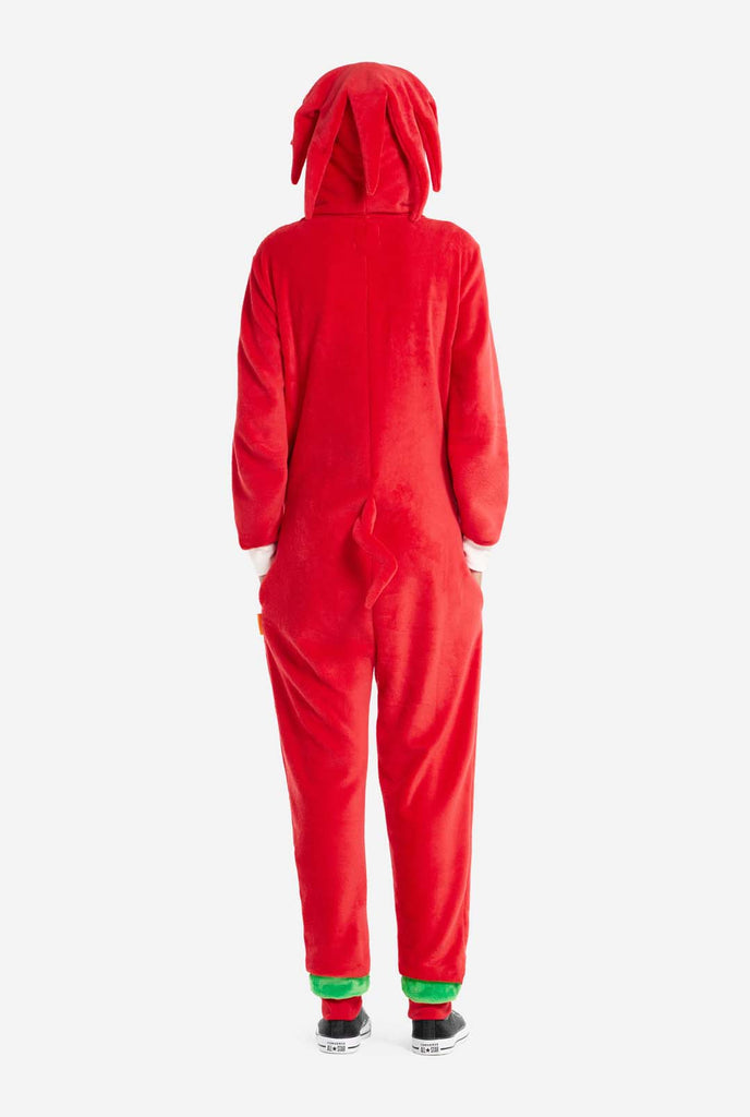 A woman wears a bright red Knuckles the Echidna adult onesie, a cozy and playful outfit inspired by the famous character from the Sonic the Hedgehog series. The onesie features Knuckles' signature white chest patch and includes a hood that showcases Knuckles' face, complete with his expressive eyes and long red dreads. The woman stands with hands in the side pockets.  The outfit, made from soft fleece material, is perfect for lounging, cosplay, or themed events. View from the back.