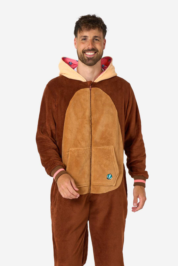 A smiling man is wearing a brown adult onesie designed as an otter. The onesie features a hood with ears, a cute face, and playful otter details, perfect for cozy lounging or costume parties. The hood is down on his back.