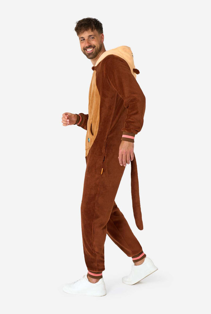 A smiling man is wearing a brown adult onesie designed as an otter. The onesie features a hood with ears, a cute face, and playful otter details, perfect for cozy lounging or costume parties. Full body image showing the side of the onesie.