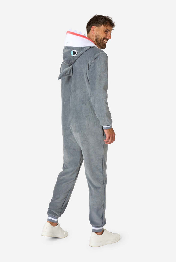 A man is wearing a grey and white shark adult onesie featuring playful hood details and cozy, soft material, perfect for lounging or costume events. Full body image from the back.