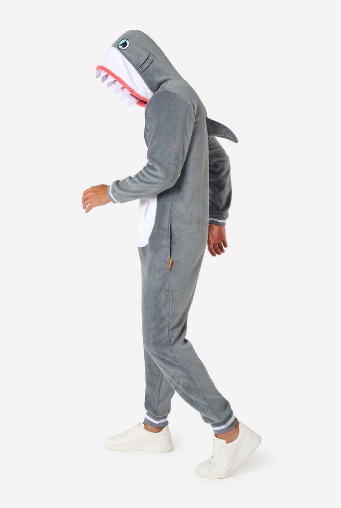 A man is wearing a grey and white shark adult onesie featuring playful hood details and cozy, soft material, perfect for lounging or costume events.  Full body image from the side.