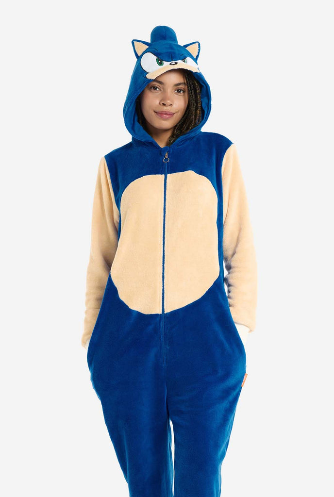 A woman is dressed in a cozy, blue Sonic the Hedgehog adult onesie, featuring soft beige patches on the front and sleeves to replicate Sonic's iconic look. The onesie has a hood designed with Sonic's recognizable face, including large green eyes, blue spikes, and pointy ears. The front zipper closure ensures a comfortable fit, and the woman stands with her hands in the onesie's side pockets. Her expression is calm and relaxed. This Sonic the Hedgehog onesie is ideal for cosplay, lounging, or themed events.