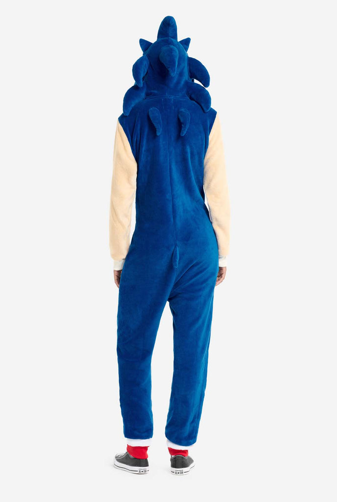 A woman is dressed in a cozy, blue Sonic the Hedgehog adult onesie, featuring soft beige sleeves to replicate Sonic's iconic look. The onesie has a hood designed with Sonic's recognizable face, including large green eyes, blue spikes, and pointy ears. This Sonic the Hedgehog onesie is ideal for cosplay, lounging, or themed events. View from the back.