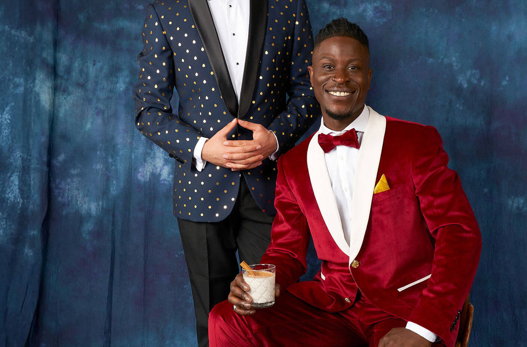 Two men in front a blue curtain wearing tuxedos. One man sitting on a chair wearing red and white velvet tuxedo. One man, standing, wearing blue and black tuxedo with golden dots.