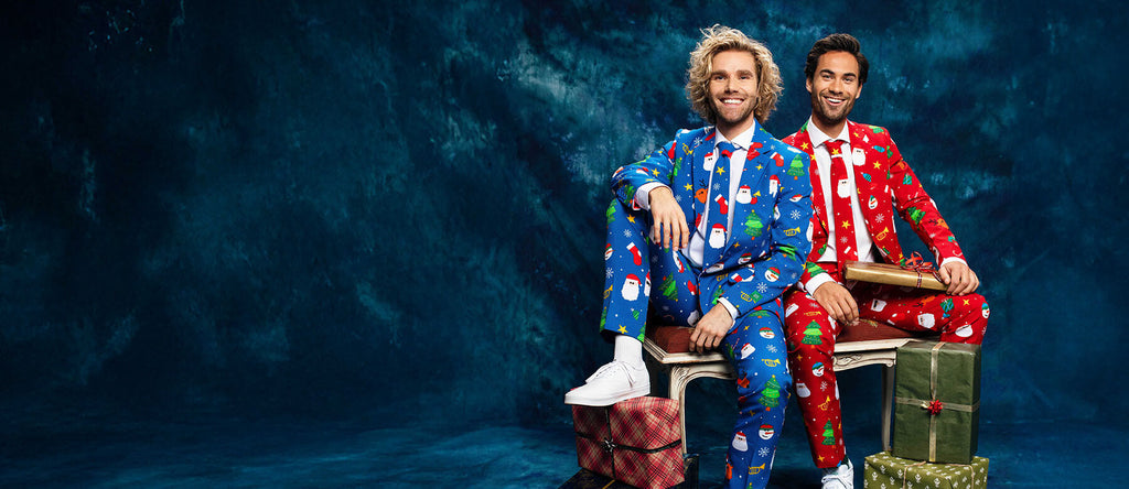 Two man sitting on a bench in fornt of a blue curtain wearing a Christmas suit. Left man wearing a blue Christmas suit and the right man wearing red Christmas suit.